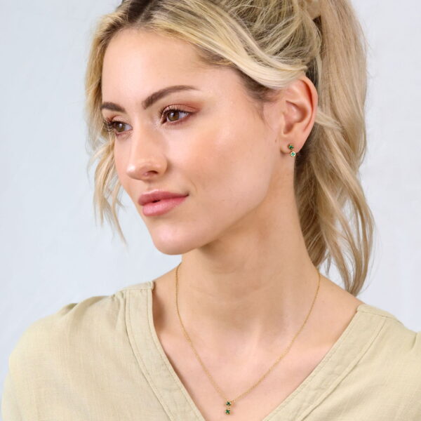 Eine Frau mit blondem Haar in einem Pferdeschwanz, die ein helles Haargummi, eine beige Bluse und kleine grüne Ohrringe trägt, modelliert die Delicate Touch Collier Mireva Halskette, während sie vor einem hellen Hintergrund zur Seite schaut.