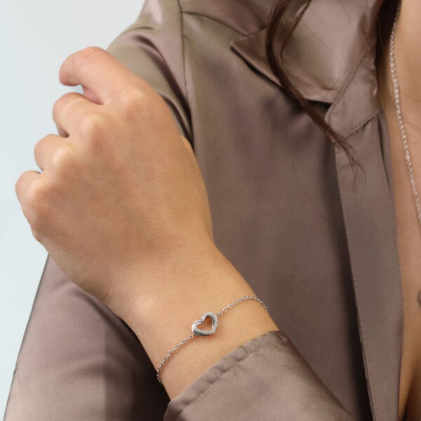 Eine Person in einer seidigen taupefarbenen Bluse trägt das Double Happiness Armband Enamored, ein zartes Silberarmband mit einem herzförmigen Anhänger, an ihrem Handgelenk. Der neutrale Hintergrund lenkt die Aufmerksamkeit auf das Schmuckstück.