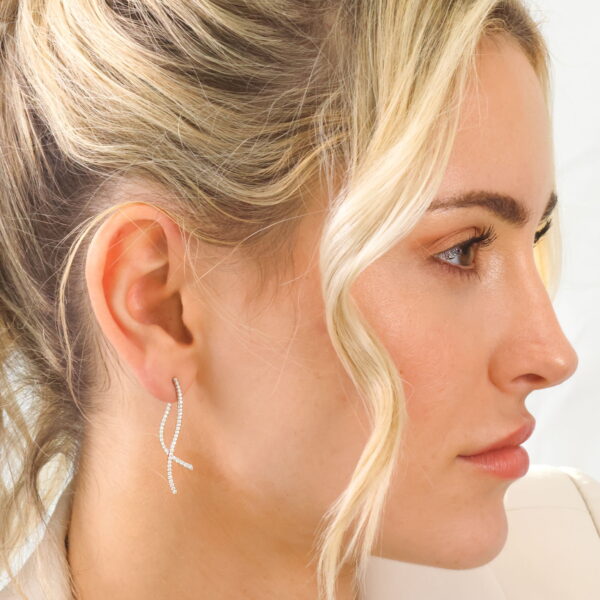 Eine Frau mit blondem Haar in einer lockeren Hochsteckfrisur trägt einen weißen Blazer und die Natural Flow Ohrringe Volura - funkelnde, geschwungene Tropfenohrringe. Sie ist im Profil vor einem hellen Hintergrund mit weichem, natürlichem Make-up abgebildet.