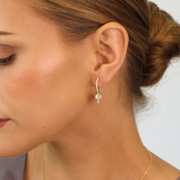 Nahaufnahme einer Frau mit heller Haut und braunem Haar in einem tiefen Dutt. Sie trägt den goldenen Reifohrring Baroque Flair Creole Naéla mit einem kleinen Anhänger und eine zarte goldene Halskette. Sie blickt nach links, wobei ihr Ohr und ihre Kieferpartie hervorgehoben werden.
