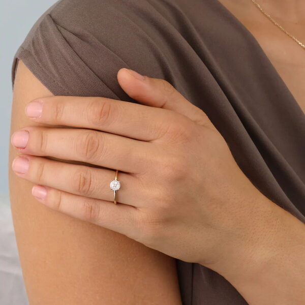 Eine Person, die eine taupefarbene Bluse trägt, stützt ihre Hand auf ihre Schulter und präsentiert den Echtgold Ring Èmera - einen Goldring mit einem runden, funkelnden Diamanten - an ihrem Ringfinger.
