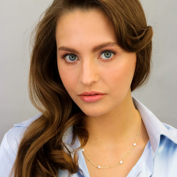 Eine Frau mit langen, gewellten braunen Haaren und blauen Augen trägt eine hellblaue Bluse und das Goldcollier Baroque Flair Adeline. Sie posiert vor einem schlichten, hellen Hintergrund mit einem neutralen Ausdruck.