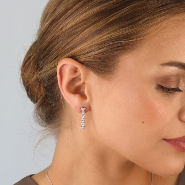 Eine Frau mit hellbraunem Haar in einem tiefen Dutt trägt Sparkly Couture Ohrringe mit Veliora-Tropfen, die mit mehreren kleinen Edelsteinen besetzt sind. Sie trägt außerdem eine zarte Halskette und blickt mit ruhiger Miene leicht zur Seite.