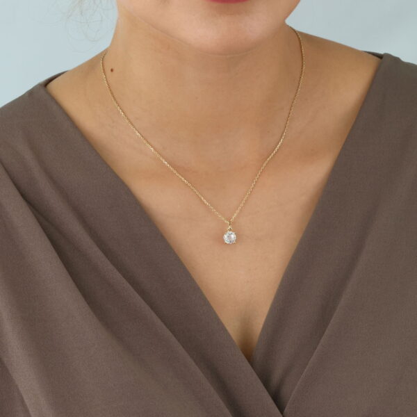 Eine Frau ist von den Schultern aufwärts vor einem hellen Hintergrund dargestellt. Sie trägt ein taupefarbenes Kleid mit V-Ausschnitt und die goldene Halskette Classic Solitaire Collier Caelia mit einem kleinen Anhänger.