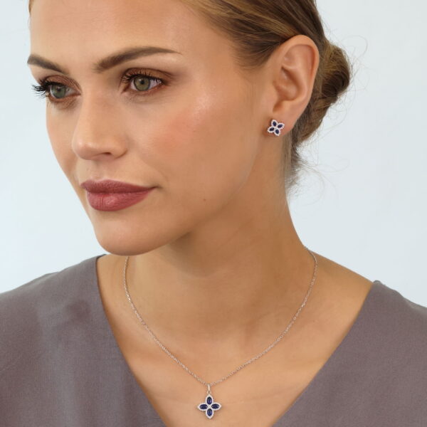 Eine Frau mit hellbraunem Haar in einem tiefen Dutt trägt eine gedämpfte lilafarbene Bluse und das Set Pretty Petals Collier Lyrienne - kleine florale Ohrringe und eine passende Anhänger-Halskette mit dunkelblauen Steinen - vor einem schlichten hellen Hintergrund.