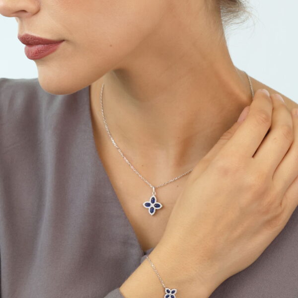 Eine Frau mit leichtem Make-up trägt eine lavendelfarbene Bluse und blickt nach unten, wobei sie ihren Hals berührt. Sie trägt das Pretty Petals Armband Lyrienne zusammen mit passenden kleeblattförmigen Ohrringen aus blauen Steinen und einer Halskette mit silbernen Konturen.
