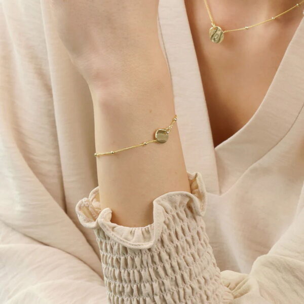 Eine Person, die ein cremefarbenes, langärmeliges Oberteil trägt, präsentiert das Modern Aztec Armband Ondelia, ein goldenes Armband mit einem kleinen runden Anhänger, gepaart mit einer passenden goldenen Halskette mit einem ähnlichen Anhänger.