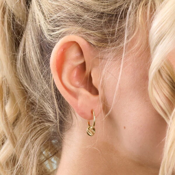 Nahaufnahme des Ohrs einer Frau, die den modernen aztekischen Creolen Ondelia trägt - einen kleinen goldenen Ohrring mit einem runden Anhänger. Ihr locker gelocktes blondes Haar umrahmt ihr Ohr.