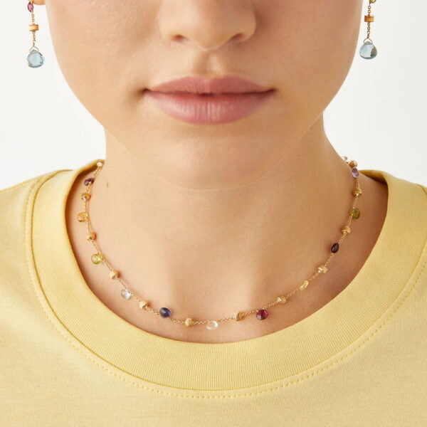 Eine Frau in einem hellgelben Hemd präsentiert die Marco Bicego Paradise Kette, eine zarte Goldkette mit kleinen mehrfarbigen Edelsteinen und passenden Ohrringen, die sich vor einem schlichten weißen Hintergrund abzeichnen.