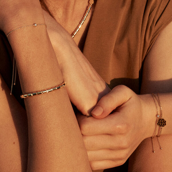 Nahaufnahme einer Person, die ein braunes Hemd und mehrere goldene Binder Classic Round 40 Armband-Armbänder an beiden Handgelenken trägt, die Arme leicht verschränkt und die Hände dicht beieinander in warmem Licht.