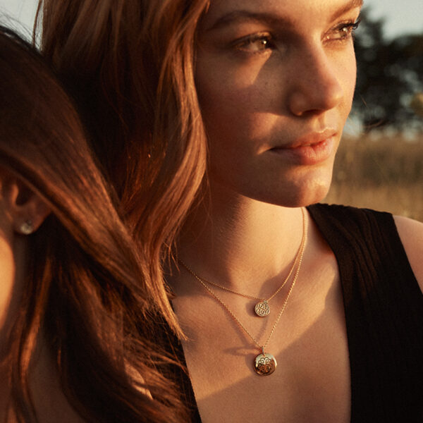 Zwei Frauen sitzen eng beieinander im Freien im warmen Sonnenlicht. Die eine, mit heller Haut und rotem Haar, trägt ein schwarzes Oberteil und die Binder Signature Kette. Sie blickt nachdenklich in den leicht verschwommenen Hintergrund, der die heitere Szene noch verstärkt.