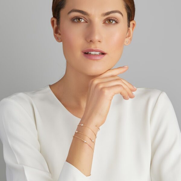 Eine Frau mit dunklem Haar in einem tiefen Dutt, einer weißen Bluse und einem zarten goldenen Yana Nesper Charmant Wickelarmband Akoya-Armband posiert nachdenklich mit der Hand am Kinn vor einem schlichten grauen Hintergrund.