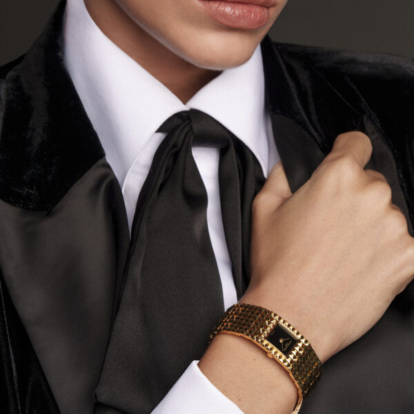Eine Person in einem schwarzen Samtanzug, weißem Hemd und schwarzer Krawatte hält ihren Kragen und präsentiert das goldene strukturierte Armband Frederique Constant Classics Manchette mit einem schwarzen Akzent. Das Bild hebt das untere Gesicht, die Hand und die Accessoires hervor.