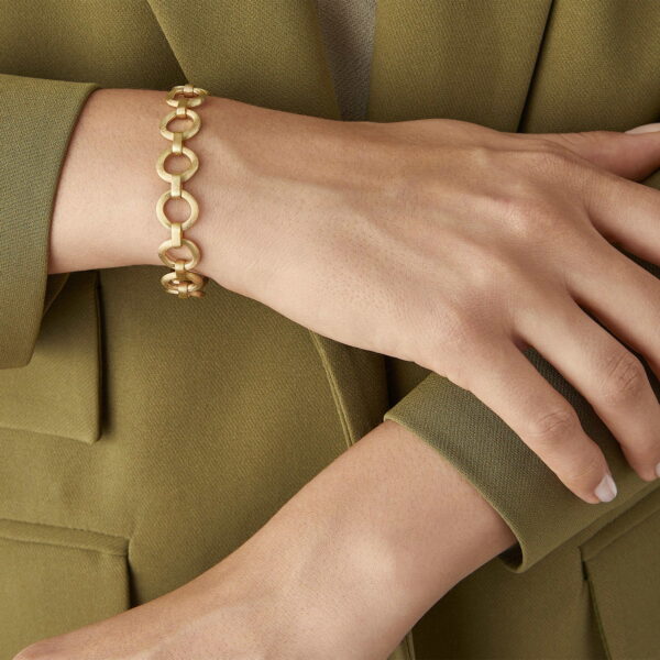 Eine Person trägt einen olivgrünen Blazer und zeigt das Marco Bicego Paradise Armband - ein goldenes Gliederarmband - an ihrem Handgelenk, wobei eine Hand sanft auf dem anderen Unterarm ruht.