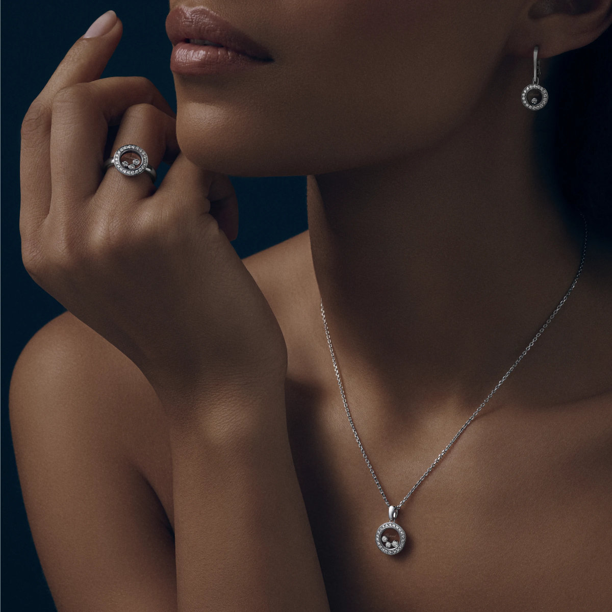 Eine Frau trägt Chopard Happy Diamonds-Schmuck - einen Ring, Ohrringe und eine Halskette - mit einem kreisförmigen Design, das einen roten Edelstein in der Mitte aufweist, der von Diamanten umgeben ist. Ihre nackte Haut hebt sich von dem dunklen Hintergrund ab.
