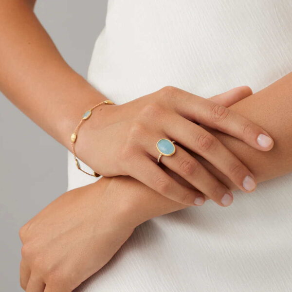 Eine Person in einem weißen, strukturierten Kleid trägt ein goldenes Armband und den Marco Bicego Siviglia Ring mit hellblauen Steinen und kreuzt sanft ihre Hände vor einem neutralen Hintergrund.