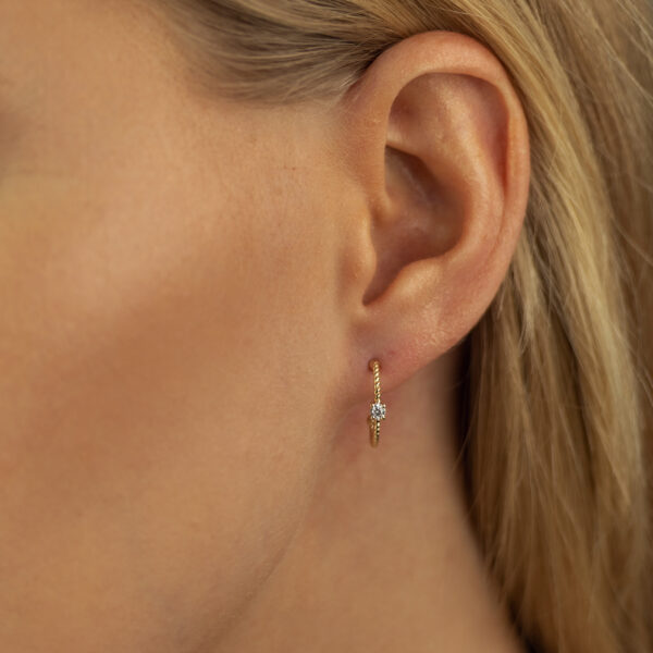 Nahaufnahme eines Frauenohrs mit glattem blondem Haar, das den Echtgold Ohrring Rayon trägt - einen zarten goldenen Ohrring mit kleinen weißen Edelsteinen, der knapp unterhalb des Ohrläppchens hängt.