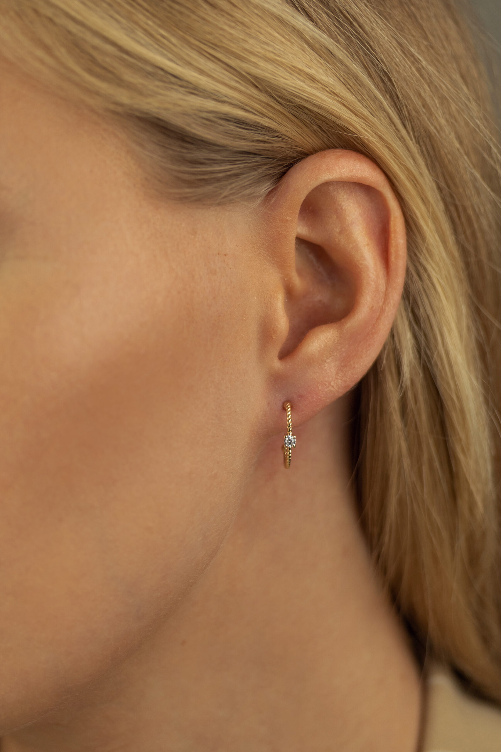 Nahaufnahme eines Frauenohrs mit glattem blondem Haar, das den Echtgold Ohrring Rayon trägt - einen zarten goldenen Ohrring mit kleinen weißen Edelsteinen, der knapp unterhalb des Ohrläppchens hängt.