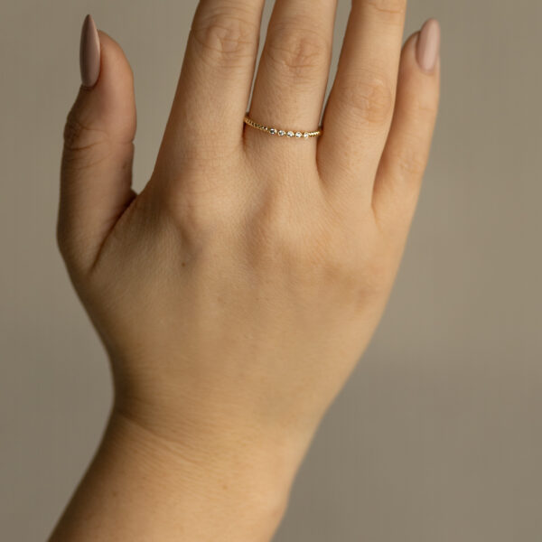 Eine Hand mit manikürten, nackten Nägeln trägt den Echtgold Ring Couronne, ein dünnes Goldband mit kleinen Edelsteinen am Mittelfinger, vor einem neutralen Hintergrund.