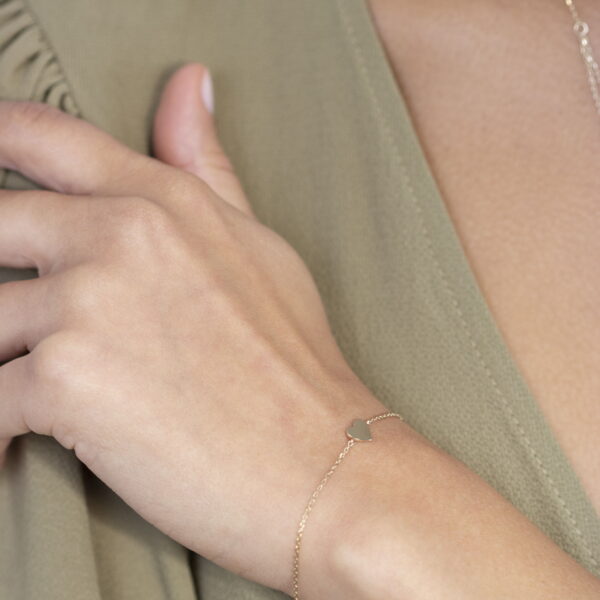 Eine Person in einem hellen olivgrünen Oberteil trägt das Echtgold Armband Loulou, ein zartes goldenes Armband mit einem kleinen Herzanhänger, und legt ihre Hand sanft auf ihre Schulter.