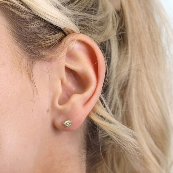 Nahaufnahme des linken Ohrs einer Frau, die Echtgold Ohrringe Rosamée trägt, einen kleinen goldenen Ohrstecker. Ihr blondes Haar ist mit einem hellen Haargummi zurückgesteckt, und lose Strähnen umrahmen sanft ihr Ohr.