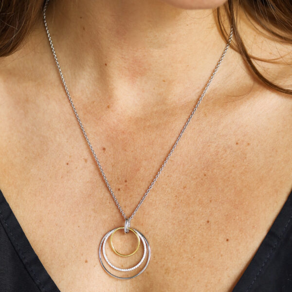 Eine Frau in einem schwarzen Oberteil mit V-Ausschnitt modelliert die Swinging Silver Halskette Bride Gold, eine silberne Halskette mit drei ineinandergreifenden kreisförmigen Anhängern und einem goldenen Ring, die sie elegant entlang ihres Schlüsselbeins drapiert.