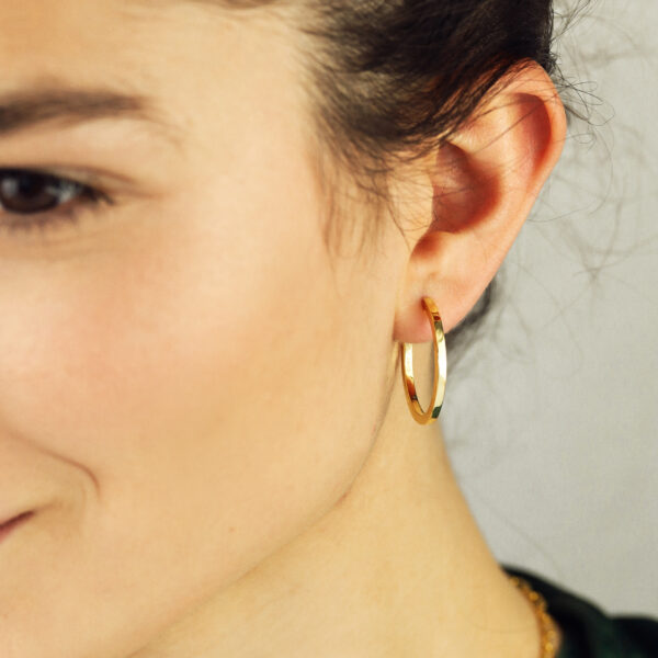 Nahaufnahme einer Frau mit heller Haut und braunem Haar in einem grünen Oberteil, die ihr Ohr mit dem Glamour Girl Creole Small Gold Hoop Earring vor einem neutralen Hintergrund präsentiert.