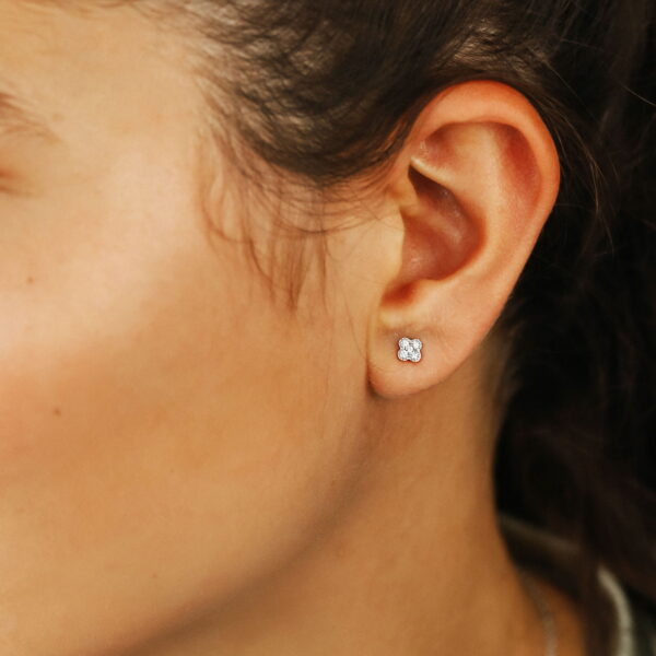 Nahaufnahme eines Frauenohrs, das mit dem Delicate Touch Ohrstecker Clover, einem kleinen, runden Ohrstecker, geschmückt ist. Ihr Haar ist zurückgekämmt und gibt den Blick auf glatte Haut, einen Teil ihres Halses und eine silberne Halskette frei.