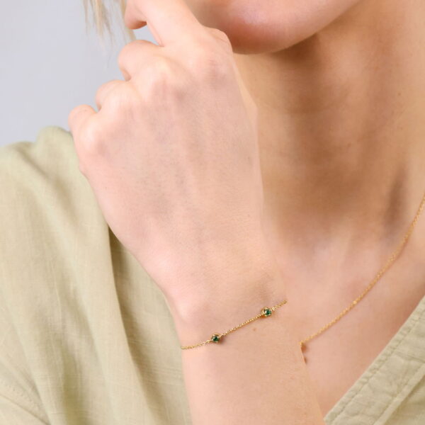 Eine Frau in einem hellgrünen Oberteil präsentiert das Delicate Touch Armband Mireva, ein goldenes Armband, das mit kleinen grünen Steinen verziert ist, zusammen mit einer passenden Halskette. Ihre Hand liegt in der Nähe ihres Kinns und ihre Lippen sind sanft gescheitelt.