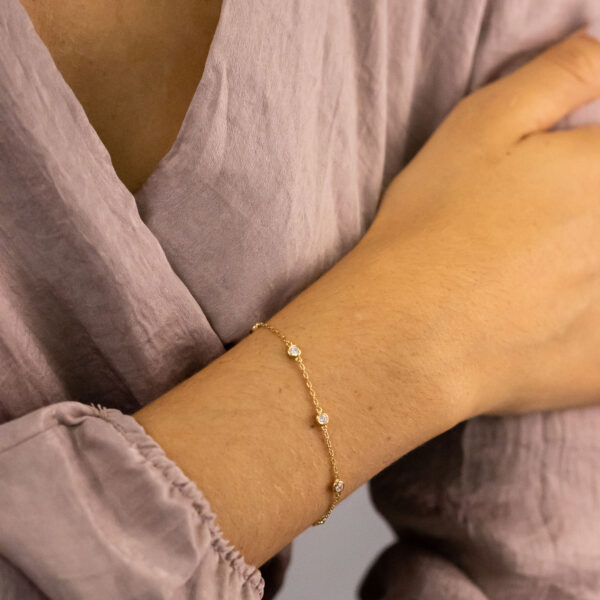 Eine Person, die eine lilafarbene Bluse trägt, kreuzt ihren Arm und trägt das Tiny Gems Armband Lynette - ein zartes Goldarmband mit kleinen runden Steinen - an ihrem Handgelenk.