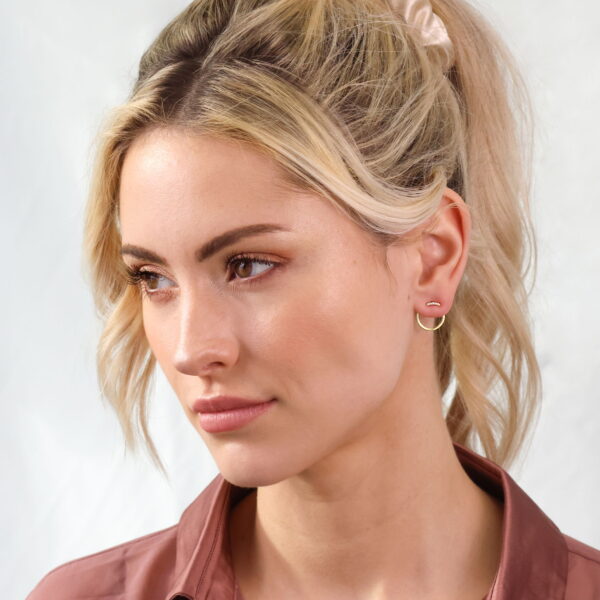 Eine Frau mit blondem Haar in einem Pferdeschwanz und cremefarbenem Haargummi, die Ohrstecker Ciréa von French Chic, goldene Ohrringe und ein lila geknöpftes Hemd trägt, schaut vor einem hellen Hintergrund nachdenklich zur Seite.