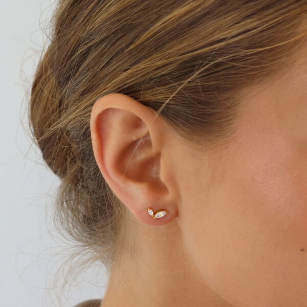 Nahaufnahme des Ohrs einer Frau, die den Sparkly Couture Ohrstecker Katelyn trägt, einen kleinen blattförmigen Ohrstecker aus Gold und Diamanten. Ihr braunes Haar trägt sie zu einem tiefen Dutt, der durch ein helles Oberteil und einen neutralen Hintergrund ergänzt wird.