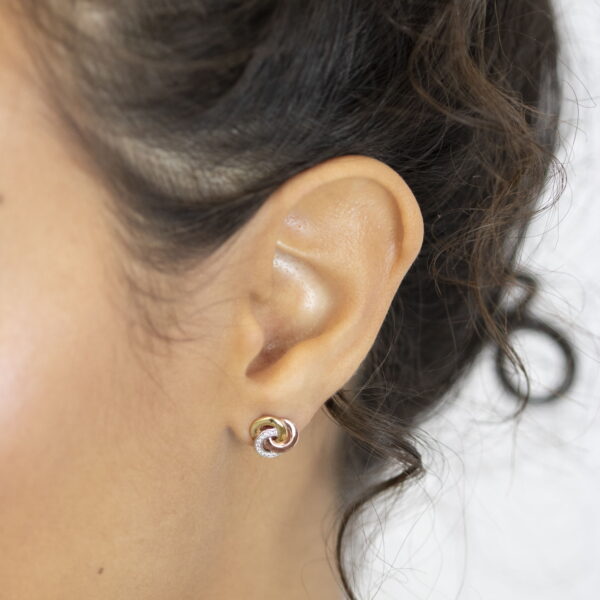 Nahaufnahme eines Frauenohrs mit dunklem, lockigem Haar, das den Echtgold Ohrstecker Noyer - kleine knotenförmige Ohrstecker in Gold-, Rosa- und Silbertönen - vor einem schlichten weißen Hintergrund trägt.