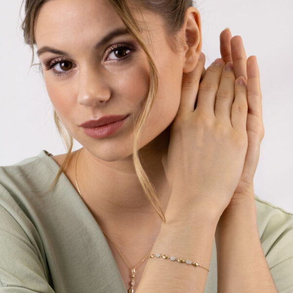 Eine Frau mit hellbraunem Haar in einer lockeren Hochsteckfrisur, die ein hellgrünes Oberteil trägt, modelliert das Echtgold Armband affidée und Goldschmuck und berührt sanft ihre Hände in der Nähe ihres Gesichts vor einem schlichten, hellen Hintergrund.