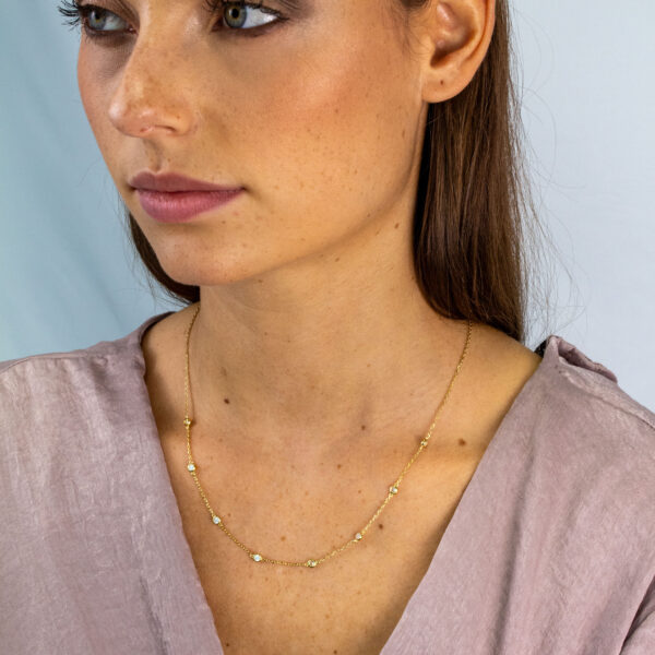 Eine Frau mit glattem braunem Haar und leichtem Make-up trägt ein mauvefarbenes strukturiertes Oberteil und das Tiny Gems Collier Lynette, eine zarte goldene Halskette mit kleinen Perlen. Sie blickt zur Seite vor einem hellen, neutralen Hintergrund.