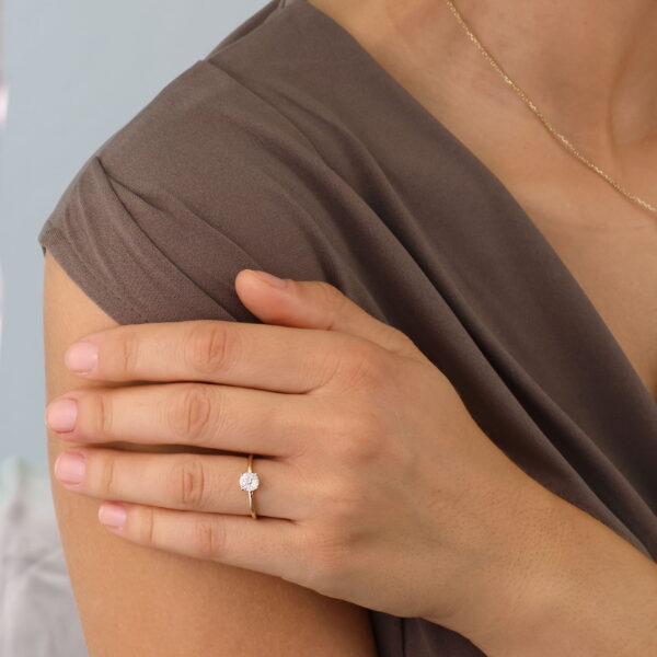 Eine Frau in einem taupefarbenen Kleid und einer zarten goldenen Halskette stützt ihre Hand auf ihre Schulter und zeigt den Classic Solitaire Ring Caelia - einen goldenen Ring mit einem kleinen runden Diamanten an ihrem Ringfinger.