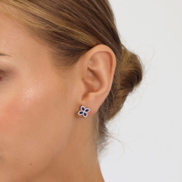 Nahaufnahme eines Frauenohrs mit Pretty Petals Ohrstecker Lyrienne, zarten blumenförmigen Ohrsteckern mit blauen und weißen Steinen. Sie trägt ihr Haar zu einem tiefen Dutt vor einem schlichten weißen Hintergrund.