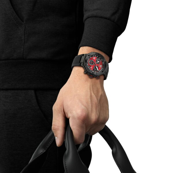 Eine Person in Schwarz hält ein schwarzes Armband und zeigt die Tissot T-Race Motogp 2026 45MM Limited Edition Armbanduhr mit rotem und schwarzem Zifferblatt. Der Hintergrund ist einfarbig weiß.