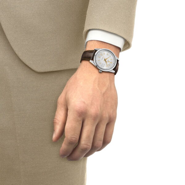 Eine Person in einem beigen Anzug und weißem Hemd präsentiert eine Tissot Visodate 39MM mit silbernem Gehäuse, braunem Lederarmband und weißem Zifferblatt an ihrem linken Handgelenk.