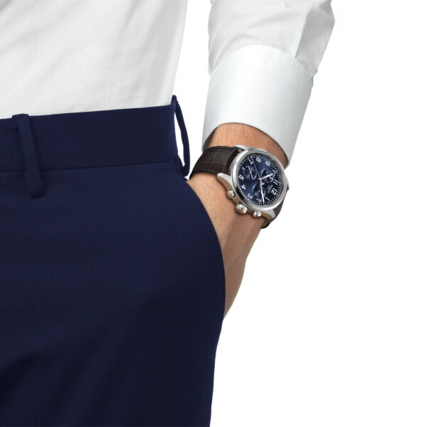 Eine Person in marineblauer Anzughose und weißem Hemd hat die linke Hand in der Tasche und zeigt eine Tissot Chrono L 42MM Armbanduhr mit schwarzem Lederband und blauem Zifferblatt.