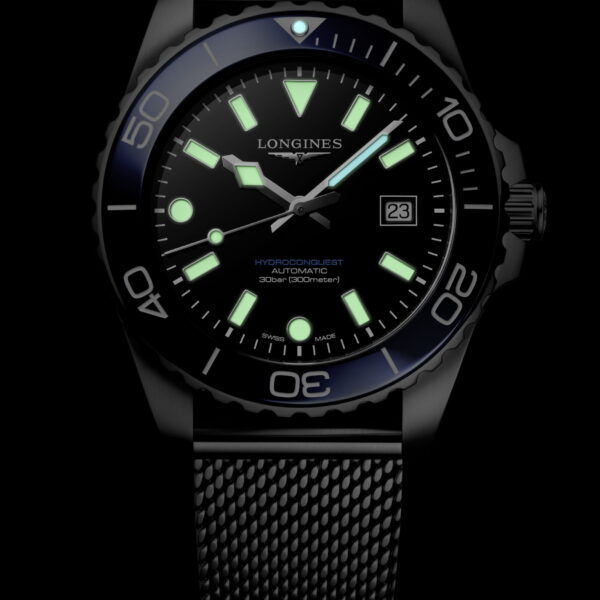 Die Longines HydroConquest, eine schwarze Taucheruhr mit Netzarmband, grünen Leuchtmarkierungen und -zeigern, Datumsanzeige bei 3 Uhr und einseitig drehbarer Lünette, wird vor einem schwarzen Hintergrund präsentiert.