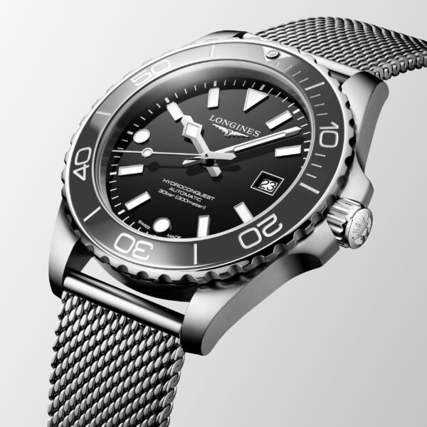 Eine Nahaufnahme der Longines HydroConquest zeigt ein schwarzes Zifferblatt, auffällige weiße Markierungen, eine drehbare Lünette und ein Edelstahlarmband aus Mesh vor einem hellgrauen Hintergrund.