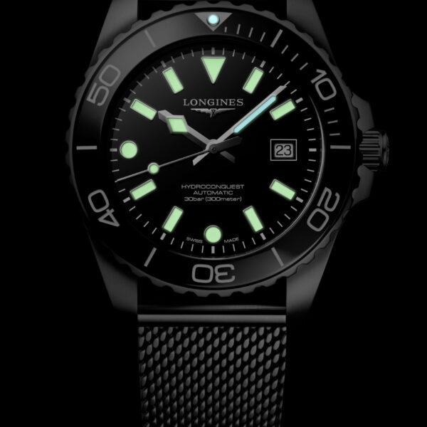 Die Longines HydroConquest Armbanduhr verfügt über ein schwarzes Zifferblatt, ein Netzarmband, grün leuchtende Markierungen, Zeiger und Lünettenakzente sowie eine Datumsanzeige bei 3 Uhr - alles vor einem markanten schwarzen Hintergrund.