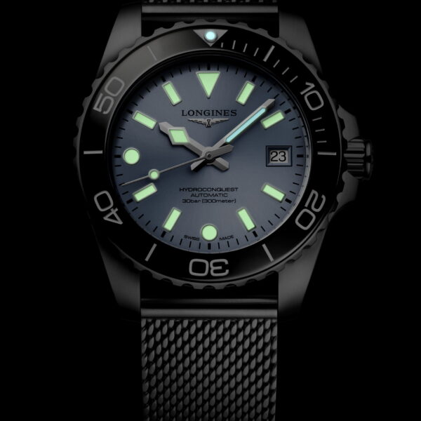 Die Longines HydroConquest ist eine schwarze Taucheruhr mit einem strukturierten Armband, blau-grün leuchtenden Indexen und Zeigern, einer Datumsanzeige und einer Lünette mit fetten Ziffern bei 15, 30 und 45 Minuten auf dunklem Hintergrund.
