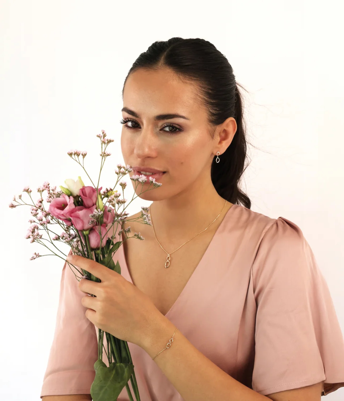Eine Frau in einem hellrosa Kleid hält einen Strauß mit rosa und weißen Blumen. Sie hat ihr langes dunkles Haar zurückgebunden und trägt zarten Goldschmuck von Nana Kay, darunter Ohrringe, eine Halskette und ein Armband, vor einem schlichten hellen Hintergrund.