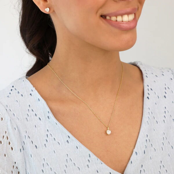 Eine Frau lächelt in einer hellblauen Ösenbluse und trägt das Classic Solitaire Collier Corin - eine goldene Halskette mit einem kleinen, runden Anhänger - und dazu passende goldene Ohrstecker, die beide mit funkelnden Steinen besetzt sind.