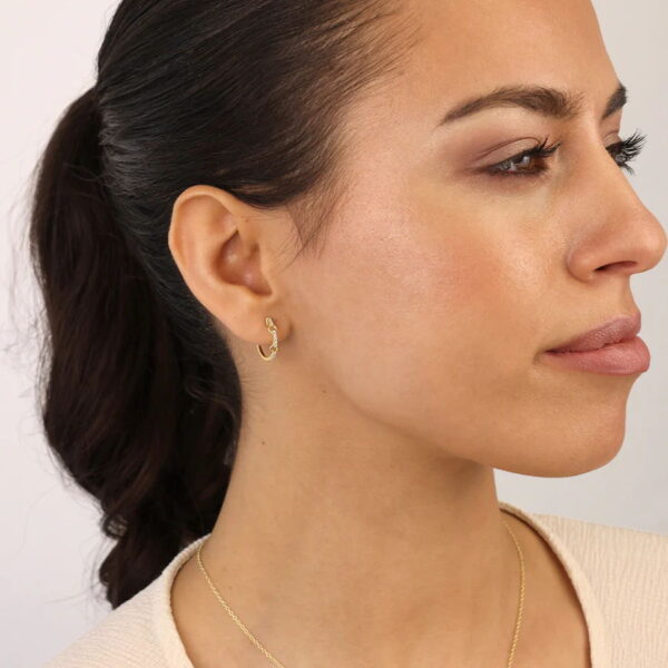 Frau mit dunklem Haar in einem Pferdeschwanz, die den goldenen Creole Aurelia-Ohrring von Solid Flair und ein helles Oberteil trägt, nach rechts schauend, mit minimalem Make-up und einem neutralen Hintergrund.
