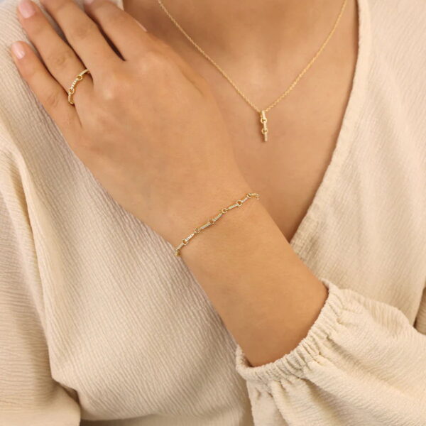 Eine Person in einem cremefarbenen, strukturierten Oberteil präsentiert das Solid Flair Armband Aurelia - zarter Goldschmuck mit kleinem Gliederdesign - zusammen mit einer passenden Halskette und einem Ring, wobei die Hand sanft ihr Schlüsselbein berührt.