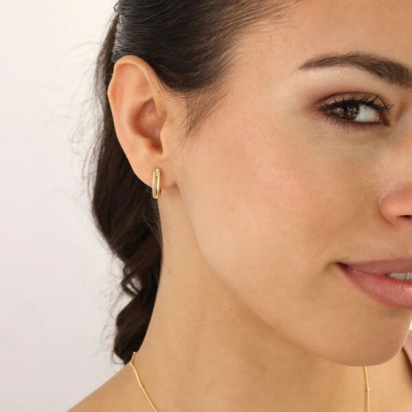 Eine Nahaufnahme einer Frau, die den Glam Rebel Creole Cordia - einen kleinen goldenen Ohrring - trägt, zeigt ihre glatte Haut und ihr minimales Make-up. Sie trägt außerdem eine zarte goldene Halskette, und ihr Haar ist zurückgesteckt, um den Ohrring zu betonen.