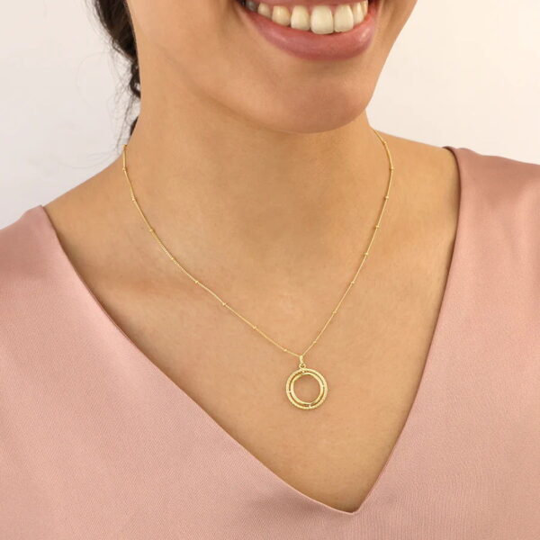 Eine Frau lächelt, während sie die Glam Rebel Collier Cordia, eine zarte goldene Halskette mit einem strukturierten runden Anhänger, zu einem hellrosa Oberteil mit V-Ausschnitt trägt.